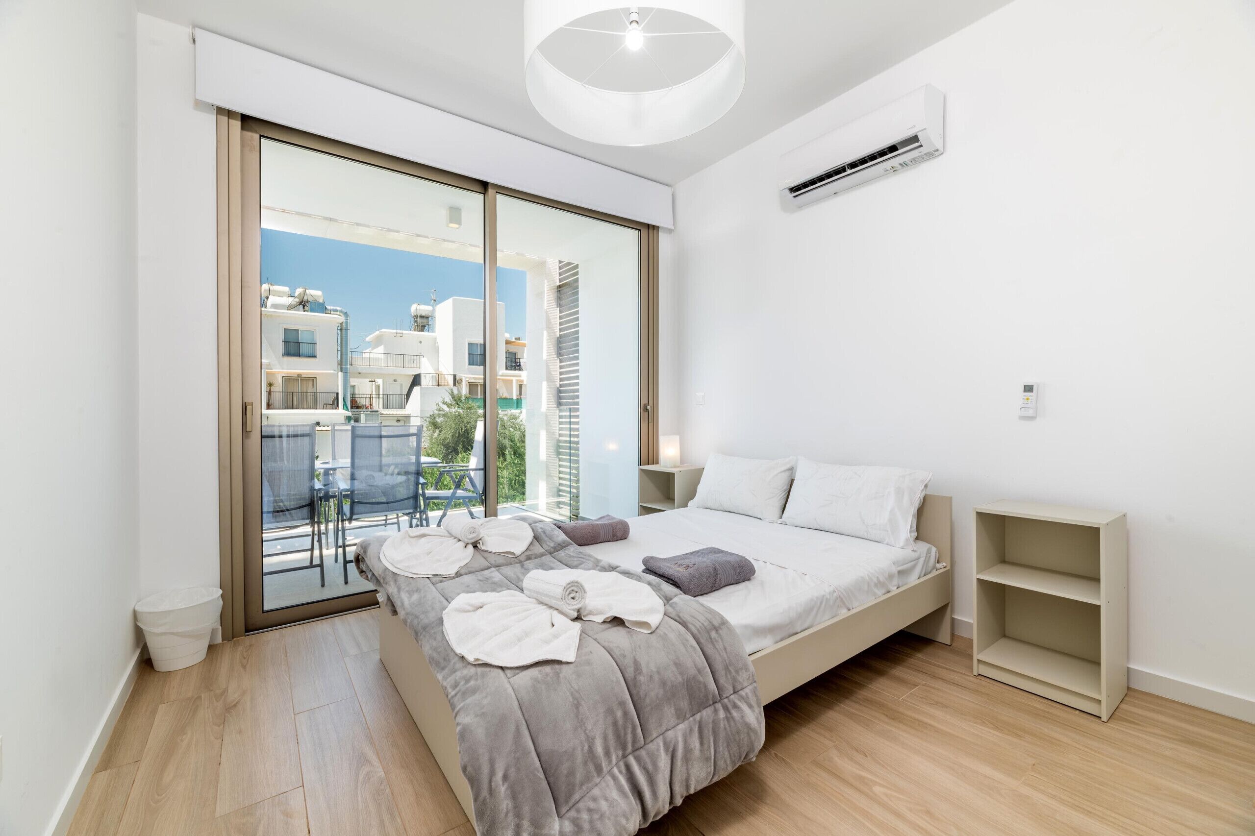 Ein modernes Schlafzimmer mit einer großen Schiebeglasdoor, die zu einem Balkon führt, einem Bett mit grauer Bettdecke, einem Nachttisch und einer kleinen Ablage.