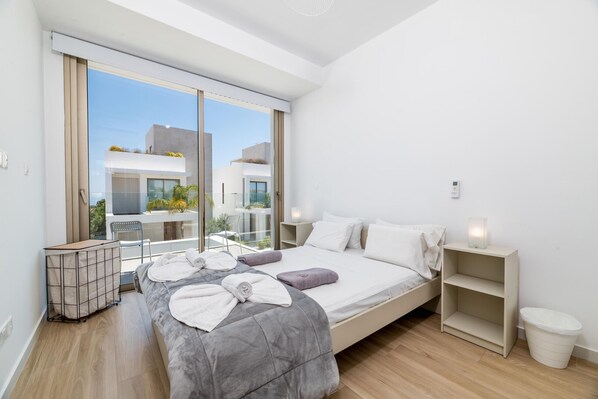 Een moderne slaapkamer met een groot bed, een nachtkastje, een raam met uitzicht op gebouwen en een balkon met een stoel en tafel.