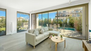 A modern living room with a beige sofa, a round wooden coffee table, and a large sliding glass door leading to a balcony with a pool and greenery.