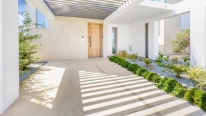 A modern building with a concrete pathway leading to a wooden door, surrounded by greenery and a pergola structure.