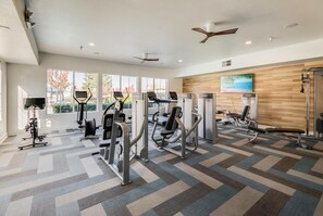 A modern gym with various exercise machines, large windows, and a wooden wall with a painting. - Lovely 1-bedroom Apartment In Great Location. (Roseville)