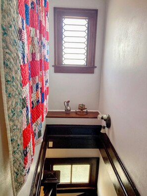A staircase with a quilt hanging on the wall, a window with a wooden frame, and a small shelf with decorative items.