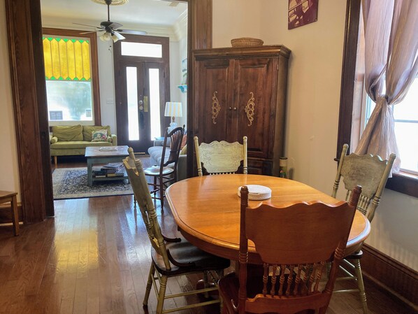 A dining area with a wooden table and chairs, a sofa in the background, and a window with curtains.