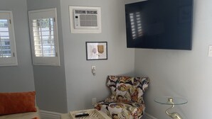 A living room with a beige sofa, a matching ottoman, a floral armchair, a glass-top coffee table, a floor lamp, a flat-screen TV, and an air conditioning unit. - Your Oasis Retreat: Pool, style, comfort, relaxation (Phoenix)