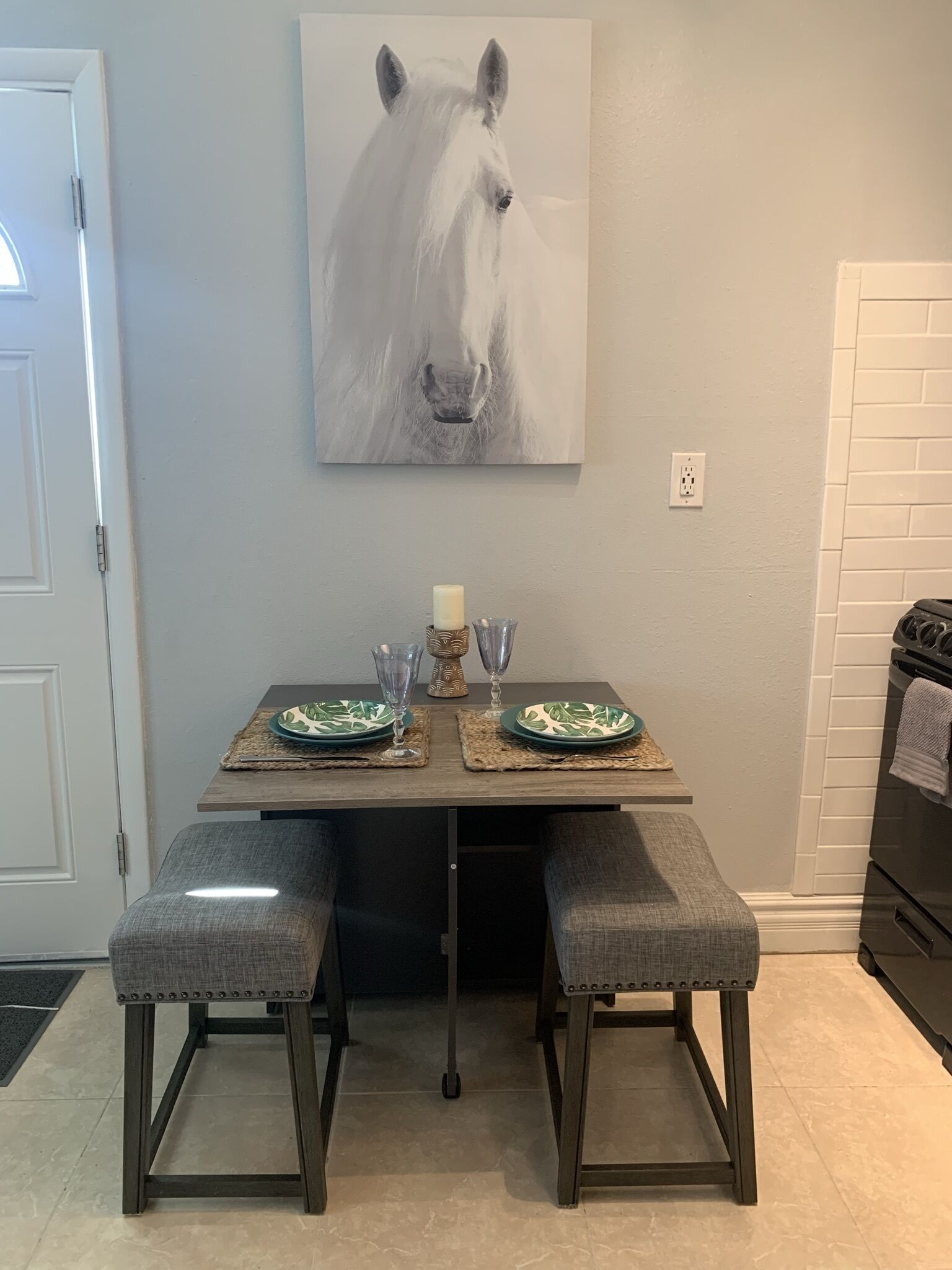 Un comedor con una mesa preparada para dos personas, con platos blancos y verdes estampados, una vela y dos vasos. Hay dos asientos tipo banca tapizados y un cuadro de un caballo en la pared.