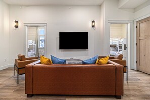 Un salon doté d’un grand téléviseur à écran plat fixé au mur, d’un canapé en cuir marron agrémenté de coussins bleus et jaunes, et de deux fauteuils.