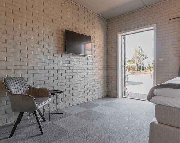 Una habitación moderna con pared de ladrillo, un televisor de pantalla plana sobre una silla, y una cama con vistas a través de una puerta de cristal.