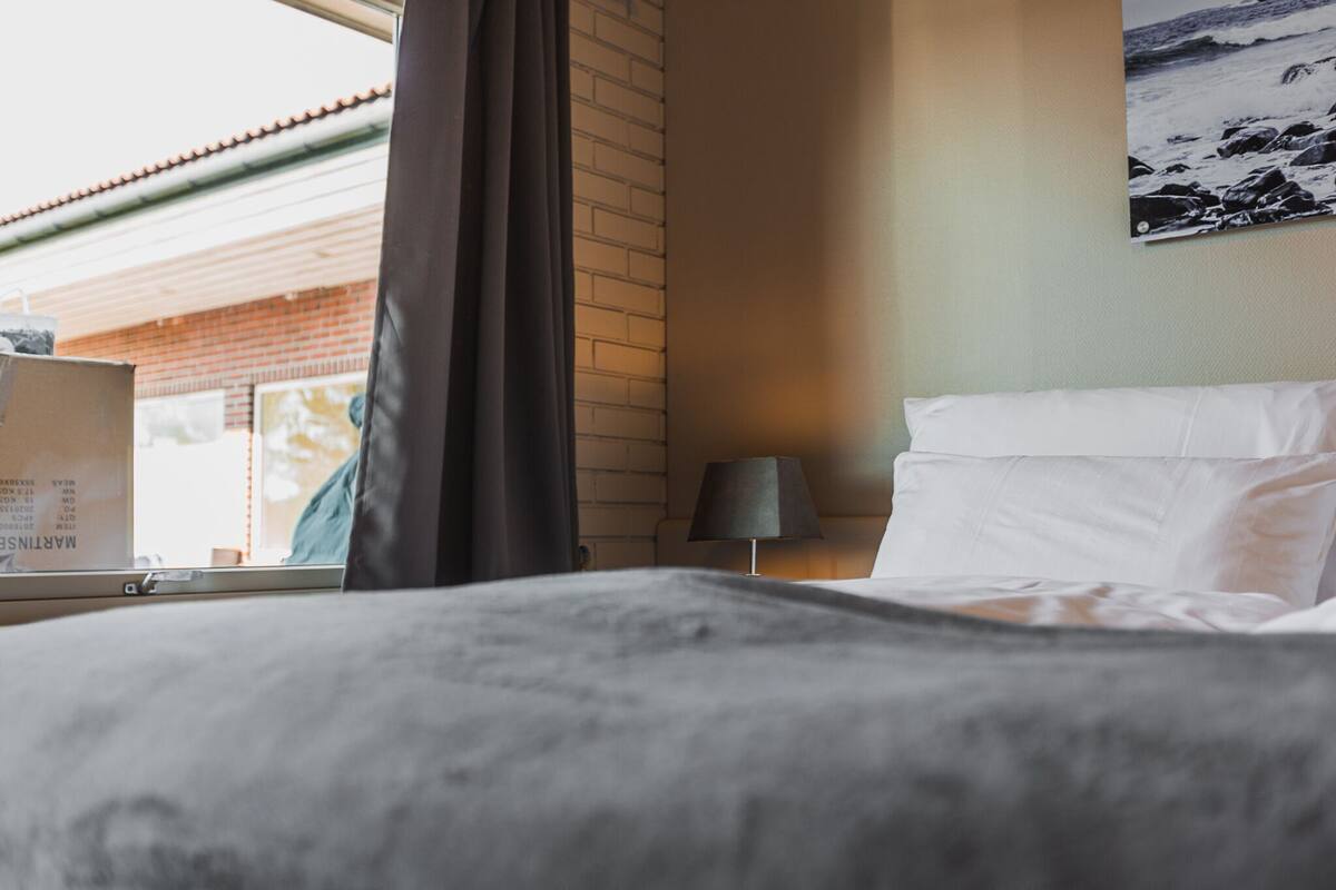 Una habitación de hotel con cama, lámpara, ventana con cortinas e imagen colgada en la pared.