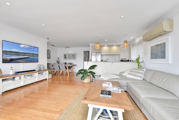 A modern living room with a flat-screen TV, a dining area with a table and chairs, a kitchen with a refrigerator and stove, and a large window with blinds.