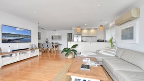 Una sala de estar moderna con una mesa de centro de madera, un sofá blanco, una zona de comedor con mesa y sillas, una cocina con armarios blancos y un televisor de pantalla plana grande en la pared.