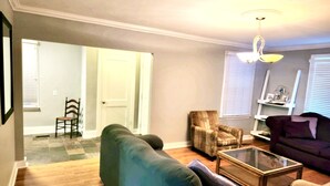 A living room with a green sofa, a wooden coffee table, a purple armchair, and a patterned armchair. There is a door leading to another room, a chair, and a shelf with items.