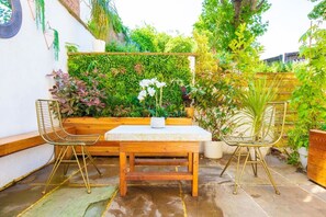 Une terrasse avec une table en bois et deux chaises, entourée d’une végétation luxuriante et d’un mur blanc.