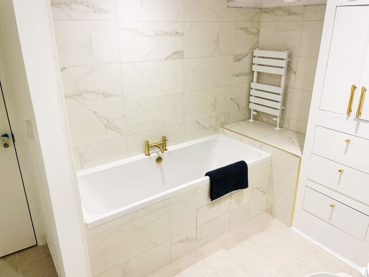 A bathroom with a white bathtub, marble tiles, a towel rack, and a white cabinet with gold handles.
