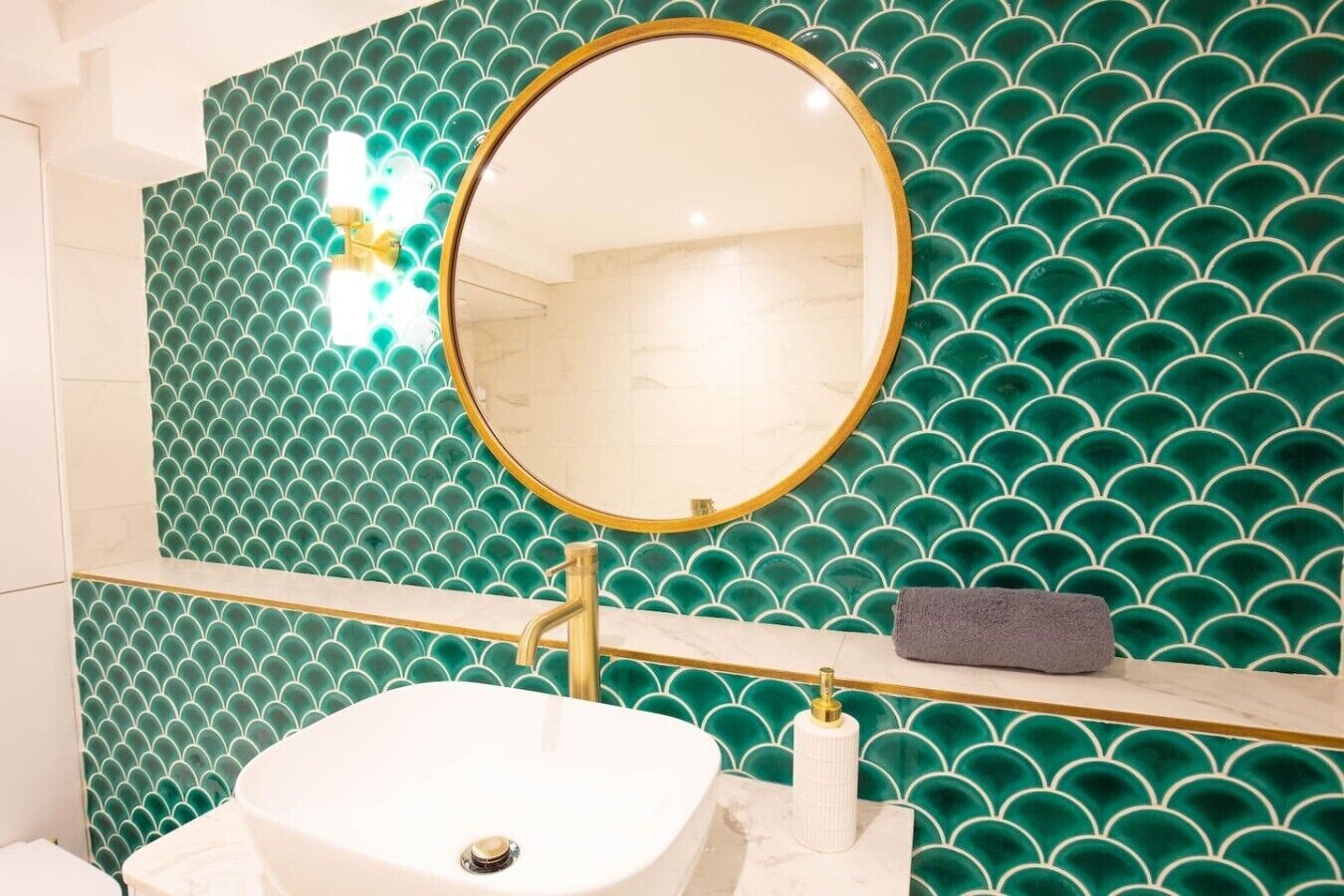 Un baño con lavamanos blanco, grifo dorado y espejo redondo con marco dorado.
