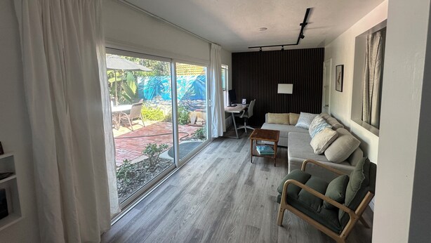 Un salón espacioso con un amplio ventanal corredera que da a un patio, un sofá, una mesita y un escritorio con una silla.