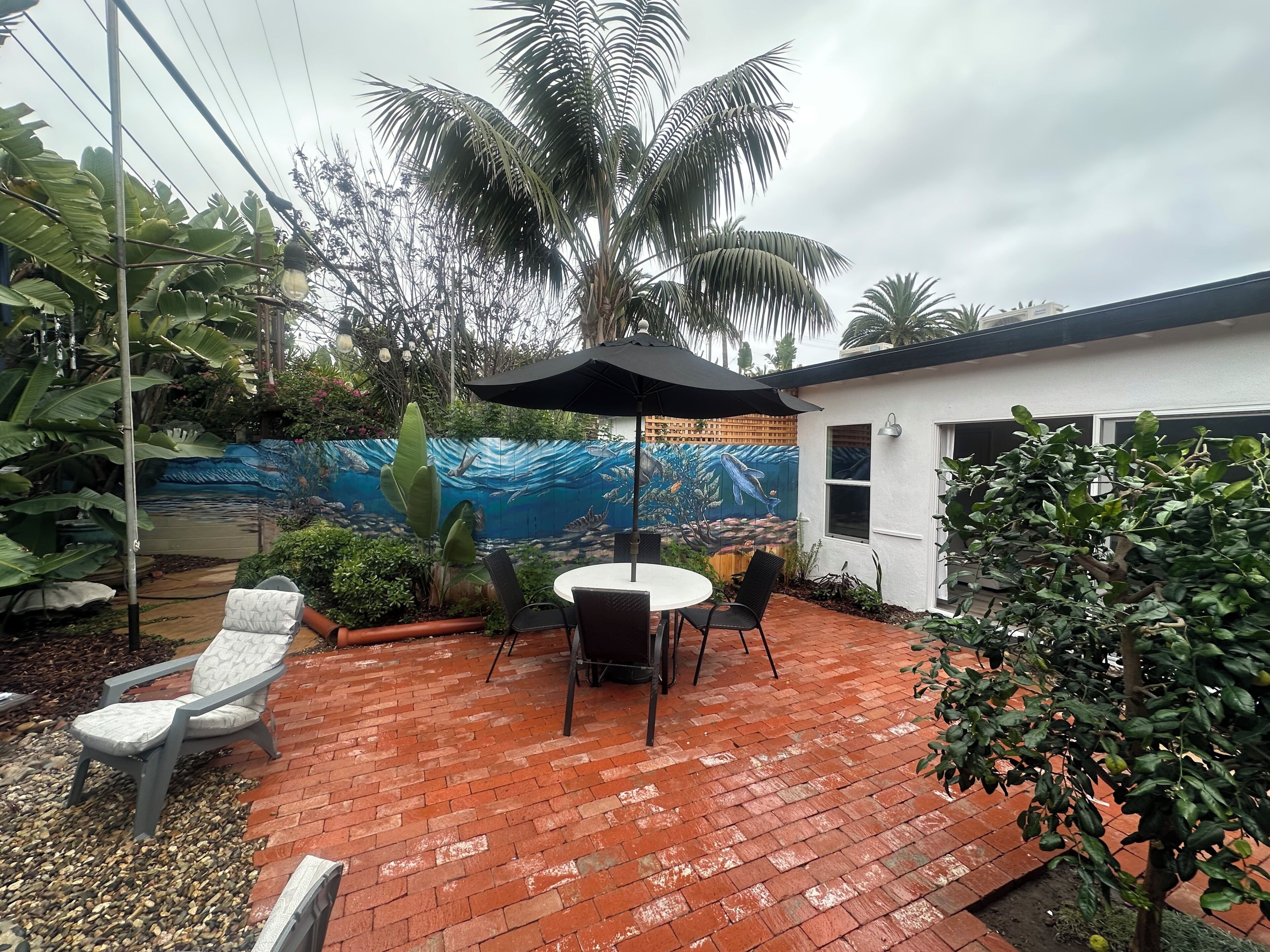 A patio with a brick floor, a round table with chairs, a black umbrella, and a mural of an underwater scene on the wall.