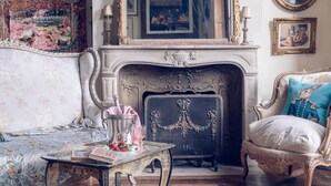 Una habitación con chimenea, una cama con ropa de cama floral, una mesita de noche con un libro y un vaso, y una silla con un cojín azul.