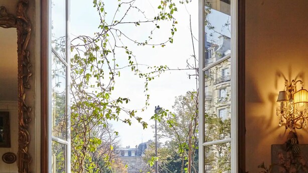 Una ventana abierta con vistas a un jardín y edificios, un banco de madera, y una mesa con libros y un jarrón de flores.