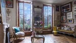 Una habitación con un ventanal grande, un sofá antiguo, una silla y una mesa con un jarrón de flores.