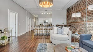 Un salon moderne avec un canapé blanc, un fauteuil bleu et une table basse. On aperçoit une cuisine en arrière-plan, avec des meubles blancs et un mur en briques.
