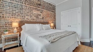 Une chambre avec un mur en briques, une tête de lit en bois, un lit blanc, deux lampes de chevet et une porte blanche.