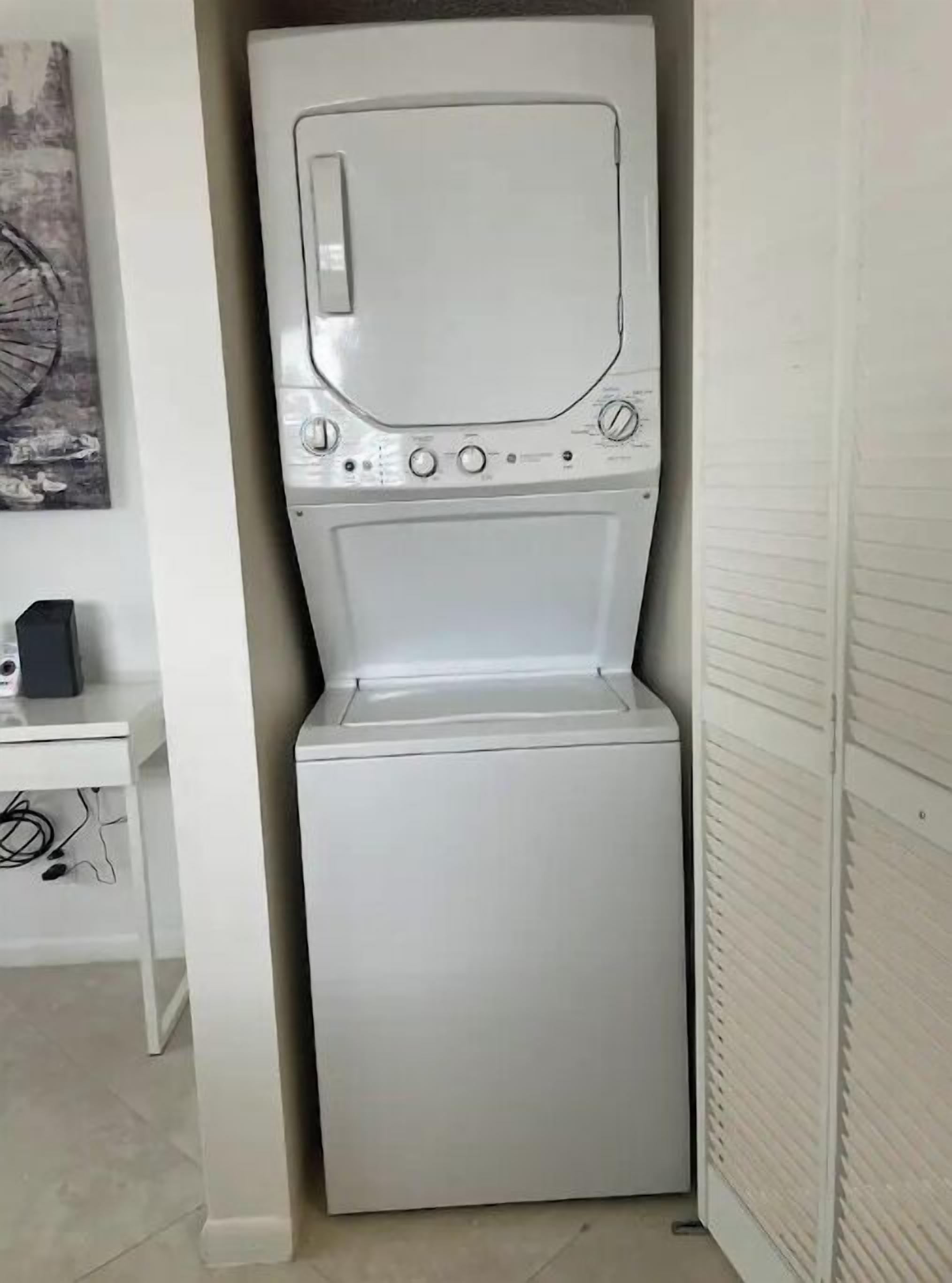 Una lavatrice a carica frontale compatta e bianca in una piccola lavanderia con un armadietto bianco e un quadro appeso al muro.