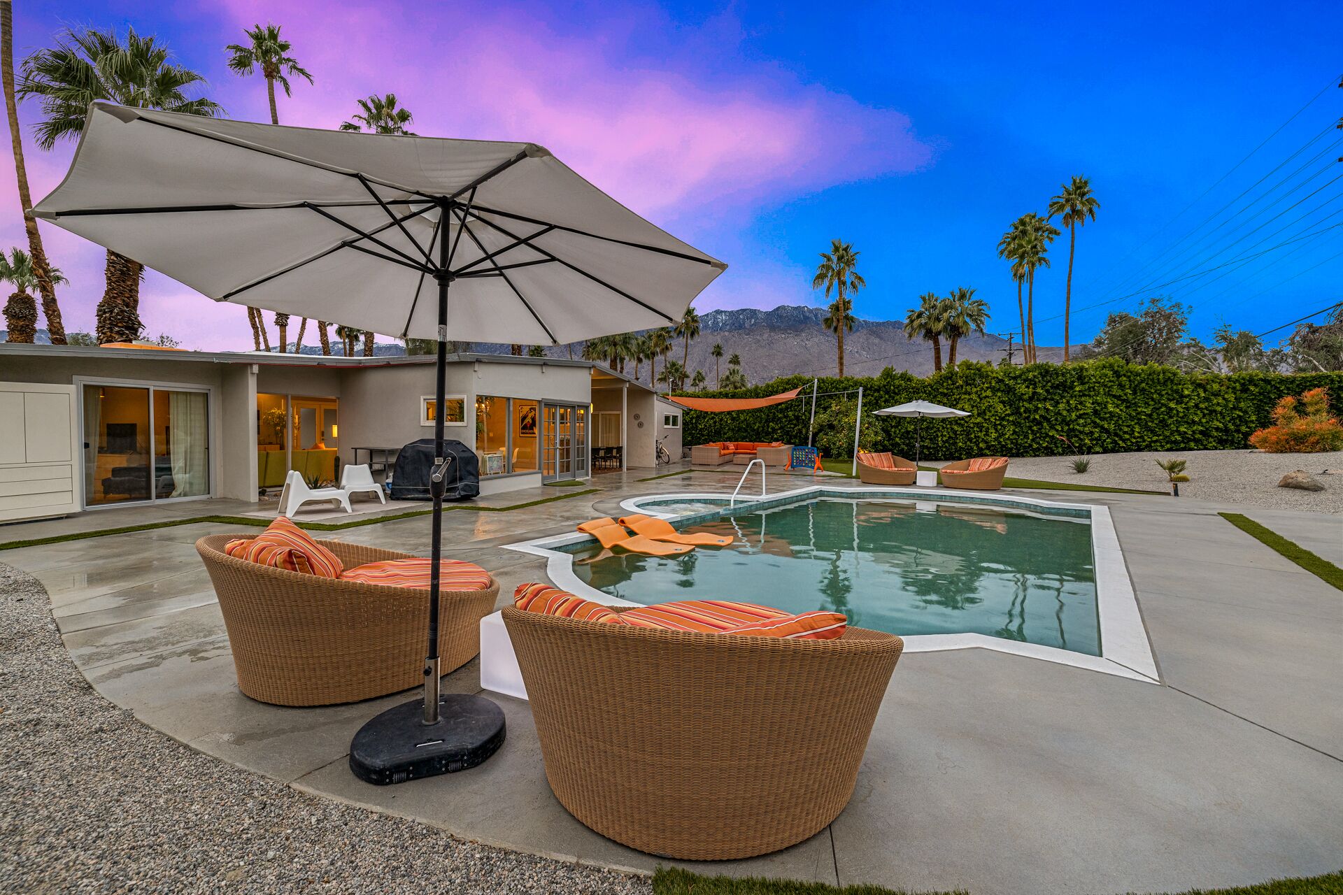 Un espace piscine extérieur moderne, doté de chaises longues et d’un parasol, entouré de palmiers et avec une chaîne de montagnes en toile de fond.