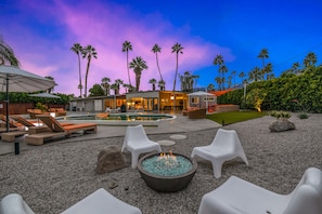 A pool area with lounge chairs, a fire pit, and a house in the background.