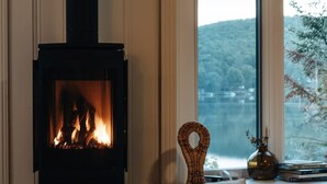 A room with a wood-burning stove, a wicker chair, and a window with a view of trees. - Lakeside Chalet, Sauna, Mont Orford & Village (Eastman)