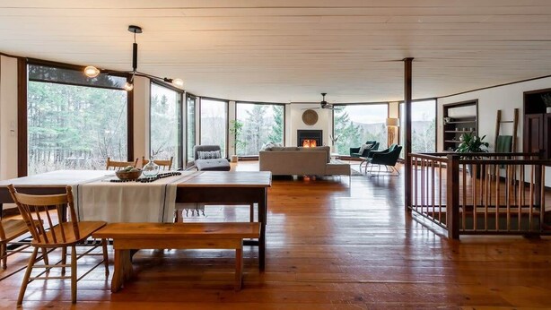 Amplia sala de estar con una mesa de comedor de madera, sillas y un sofá. Se observa una chimenea al fondo y ventanas amplias que ofrecen vistas al exterior.
