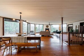 A spacious living area with a wooden dining table, chairs, and a sofa. There is a fireplace in the background and large windows offering a view of the outdoors. - L'Orford Forest Dream: Hot Tub, Fireplace & Views (Orford)