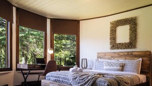 Un dormitorio con una cama de madera, un escritorio y una silla. Amplias ventanas con vista a árboles.