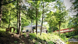 Eine Hütte mit Terrasse und Sitzgelegenheiten im Freien, umgeben von dichtem Wald und einem Bach.