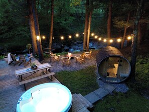 A cozy outdoor setting with a hot tub, wooden deck, chairs, and a log cabin structure. - Le 88 Howard - Spa, Sauna, River & Cute Bridge! (Mansonville)