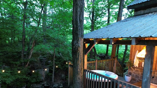 A wooden deck with a hot tub, stone fire pit, and a rustic wooden structure with a hanging light.