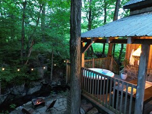 Una terraza de madera con un jacuzzi, un hogar a piedra y una estructura rústica de madera con una lámpara colgante.