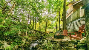 A cabin with a stone fire pit, wooden deck, and a view of a forested stream.