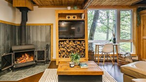 A cozy living room with a wood-burning stove, a television on a wooden shelf, and a dining area with a table and chairs.