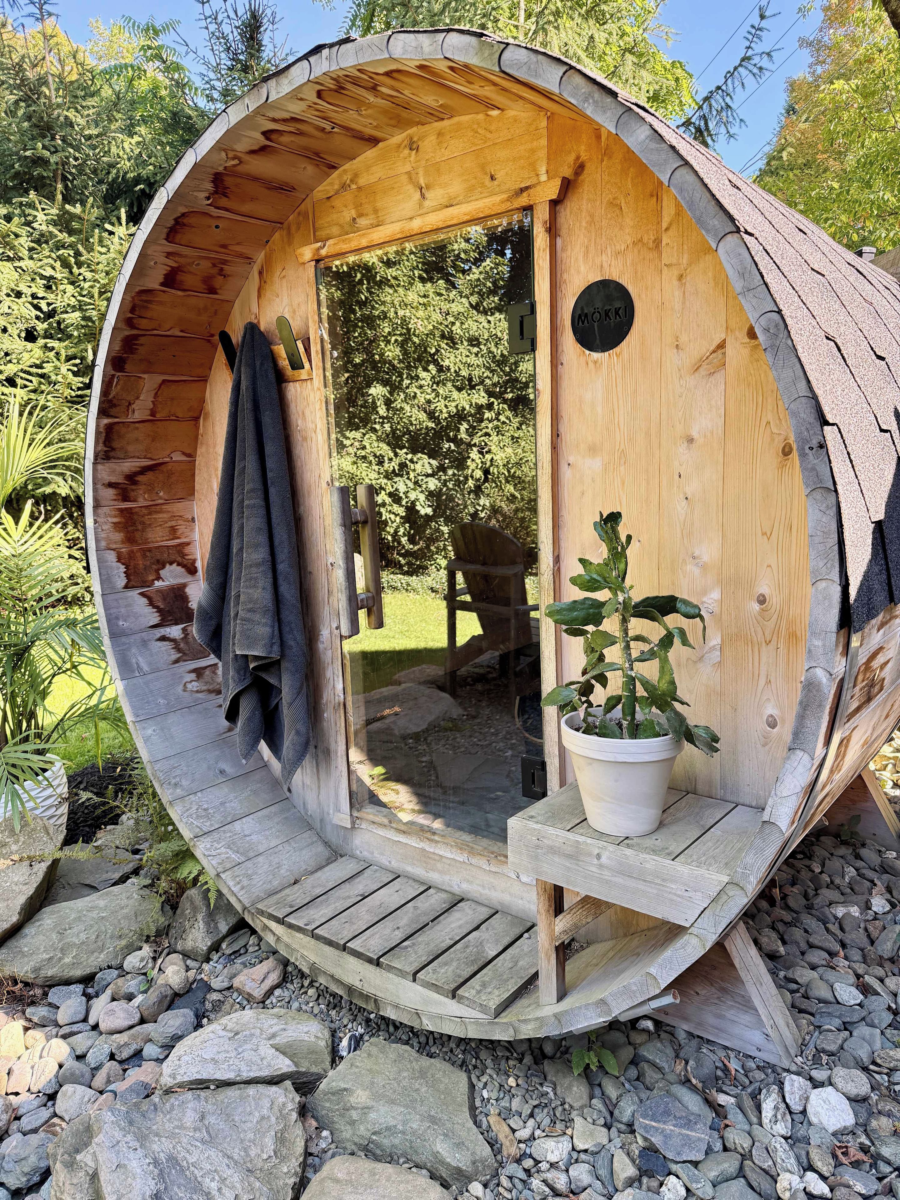 Une structure en forme de tonneau, faite de bois, avec une porte, un banc et une plante en pot à l’intérieur.