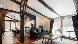 A spacious living area with a wooden floor, a black sofa, a coffee table, and a large window with curtains. - Le Cozy-St-Côme: Private Lake, Spa & Nature at Val St-Côme (Saint-Côme)