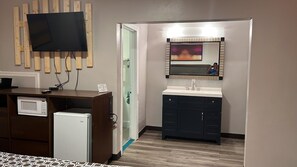 Habitación de hotel con una pared de madera, un televisor montado, un refrigerador pequeño, un microondas, un lavamanos con gabinetes oscuros y un espejo con reflejo.