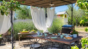 Un espace extérieur aménagé pour les repas, avec une table dressée, entourée de chaises, et un barbecue à l’arrière-plan.