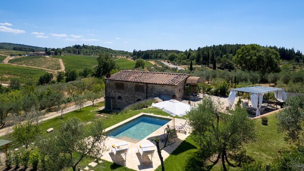 Un bâtiment en pierre avec un toit en tuiles, une piscine et un espace extérieur aménagé, couvert et entouré de verdure.
