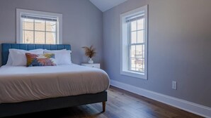 Un dormitorio con cama, dos ventanas, una mesita de noche y una planta en maceta.