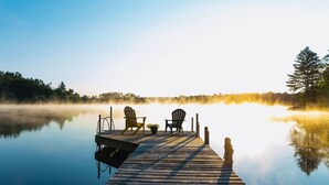 A wooden pier extending into calm water with two Adirondack chairs and a bench, surrounded by trees and a misty lake. - Waterfront Splendor in the Woods. (Florence)