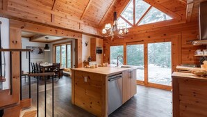 Interior de una cabaña de madera con una isla de cocina central, comedor y grandes ventanales que ofrecen vistas al bosque.
