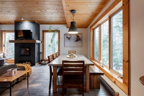 Ein gemütliches Wohnzimmer mit Kamin, Essbereich und großen Fenstern mit Blick auf den Wald.