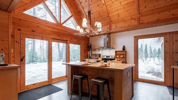 Interior de una cabaña de madera con una isla grande en la cocina, taburetes de bar y ventanas amplias que ofrecen vistas a un bosque nevado.