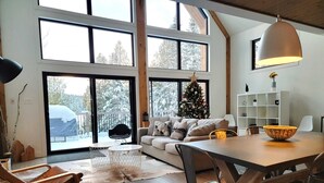 A spacious living room with a large window, a Christmas tree, a dining table, and a cozy seating area. - Nômade - Spa, private lake access & stunning views (Chertsey)