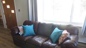 A brown leather sofa with two blue cushions and a floral cushion, positioned in a room with wooden flooring and a window. - Charming 3-bedroom farmhouse with amazing views! (Durango)